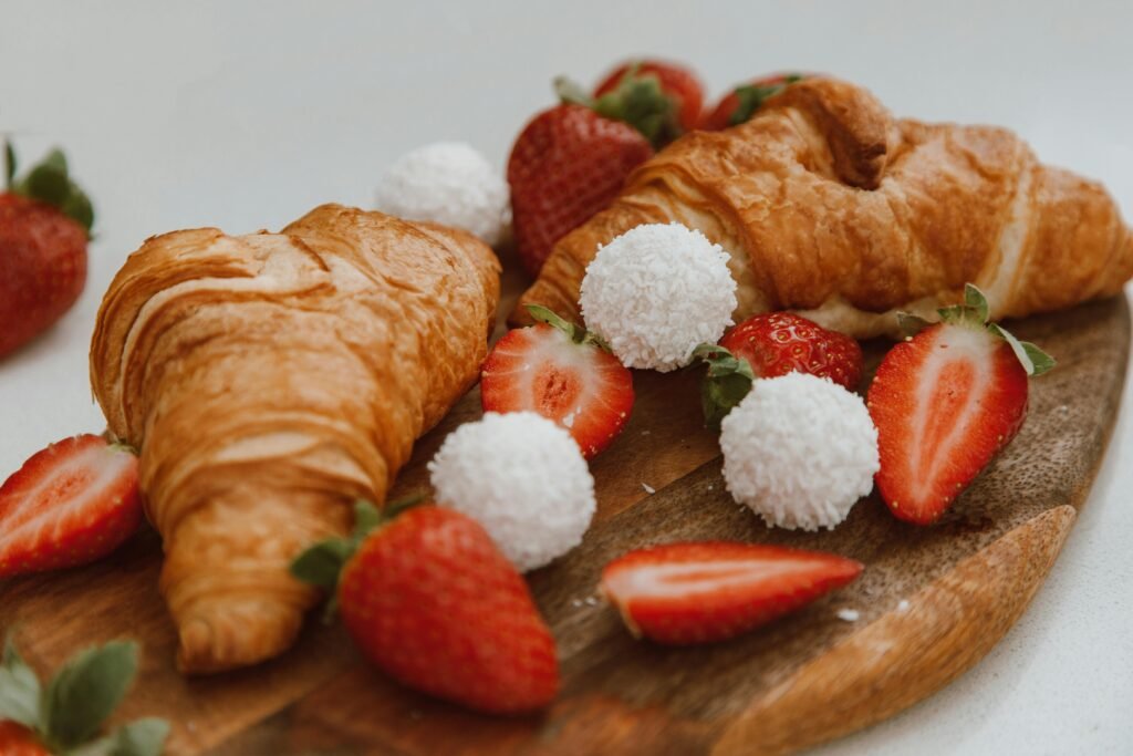 A close-up of freshly baked croissants accompanied by strawberries and coconut balls on a wooden board.